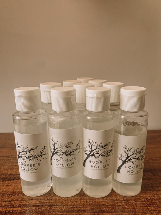 Twelve clear plastic bottles of Hooper's Hollow wood cleaner stand on a wooden surface. Each bottle features a white label with a black tree illustration and the company name "Hooper's Hollow Custom Wood Working". 
